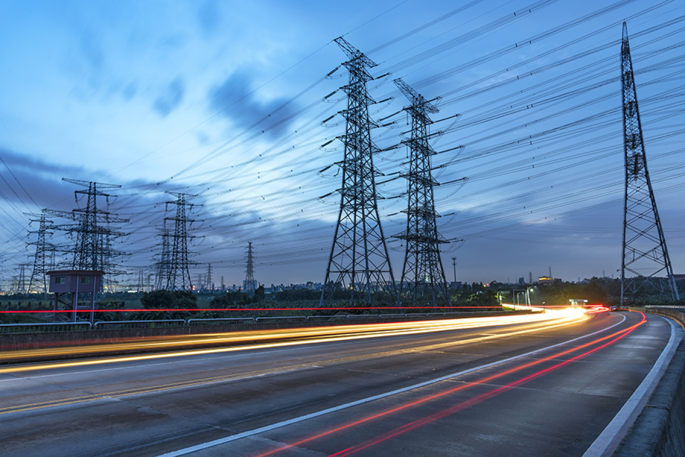 ime-lapse photo taken at dawn or dusk, showing the lights of cars driving under power transmission lines.