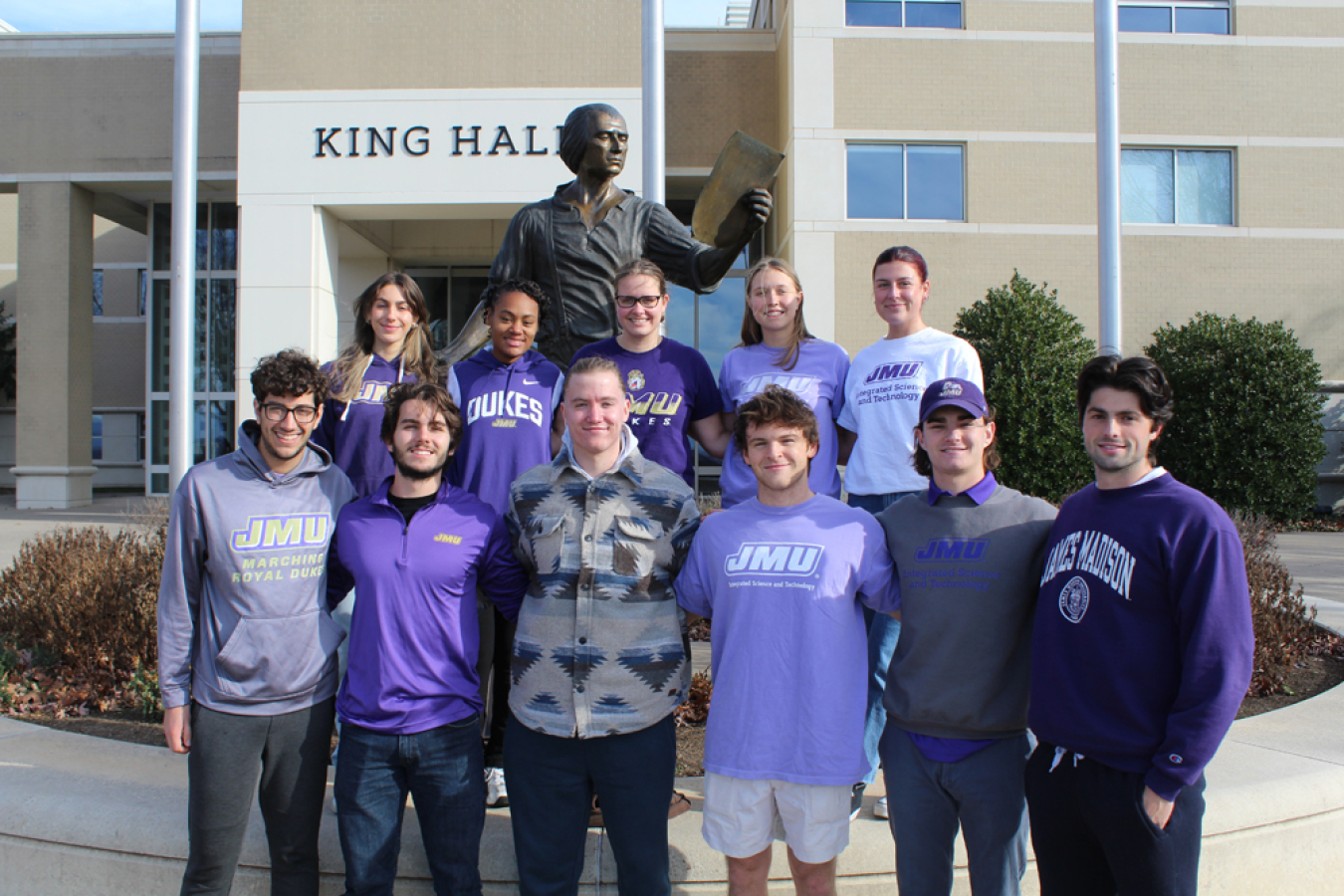 A group of people in James Madison University attire in front of a building and a statue.