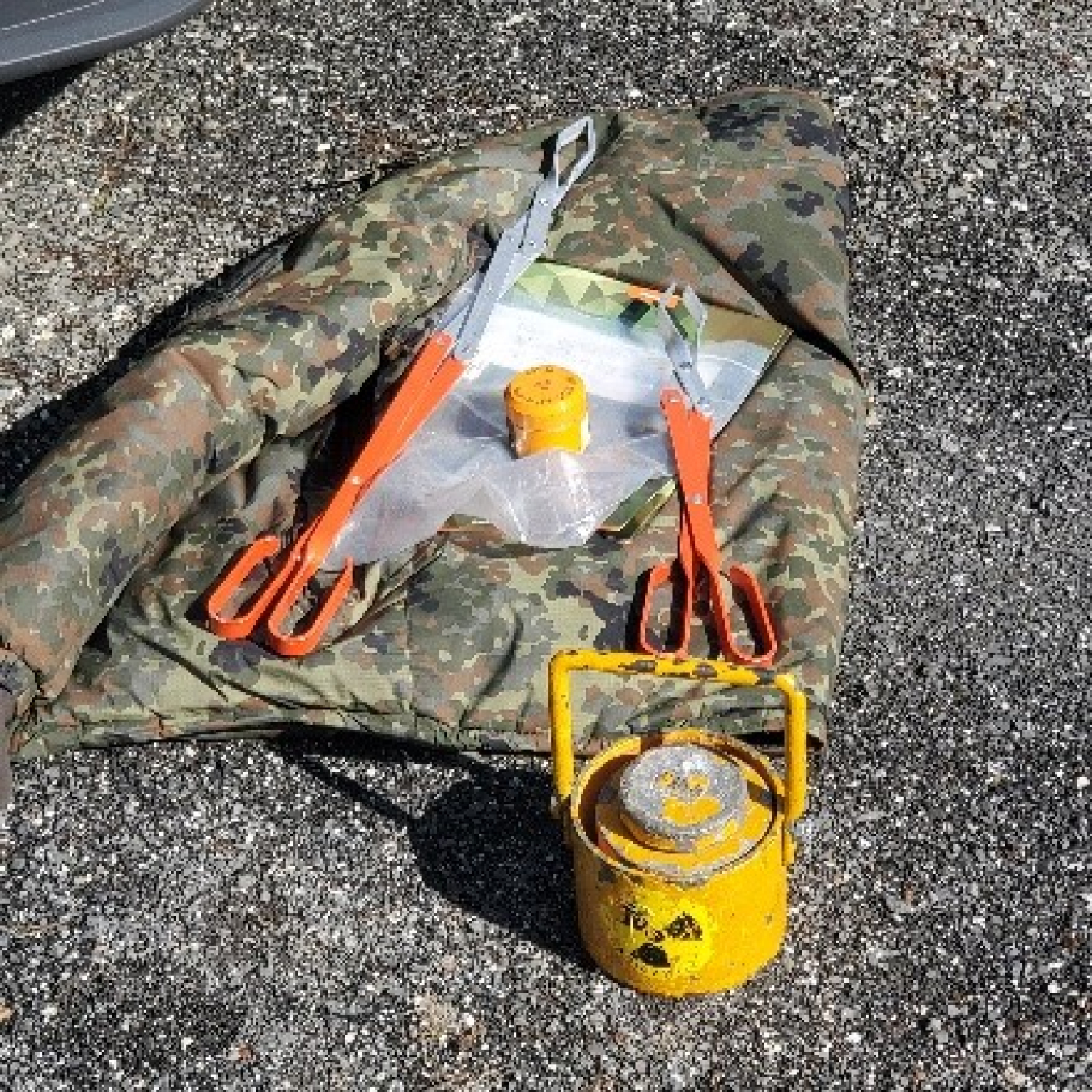 A radioactive source and a pair of industrial-type tongs lay on the ground.