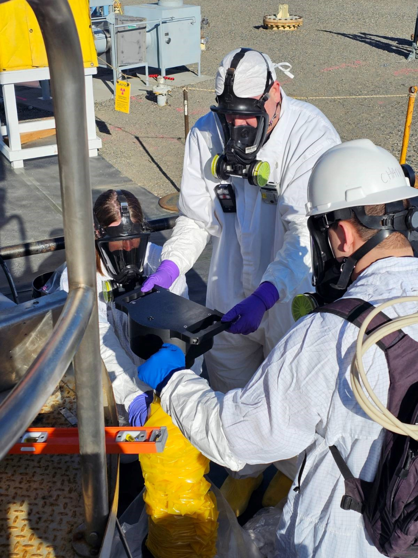 Employees in white safety suits and hard hats work outside with measurement tools