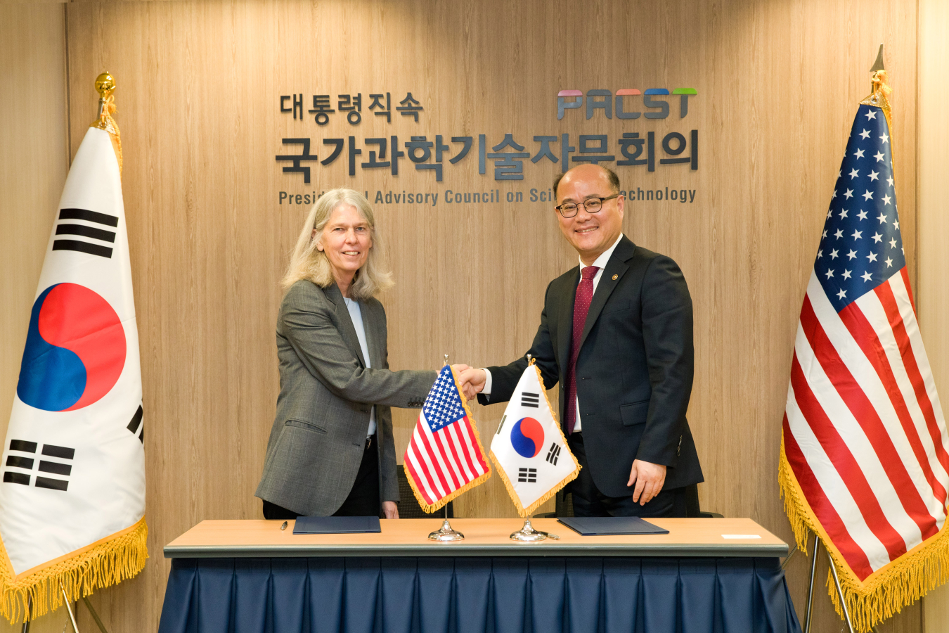 Jill Hruby and Lee Chang Yune shake hands. U.S. and Republic of Korea flags flank them.