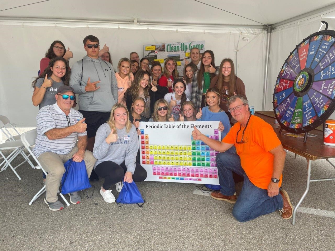 A group of people pose for a picture in a tent beside a spinning prize wheel and a periodic table poster