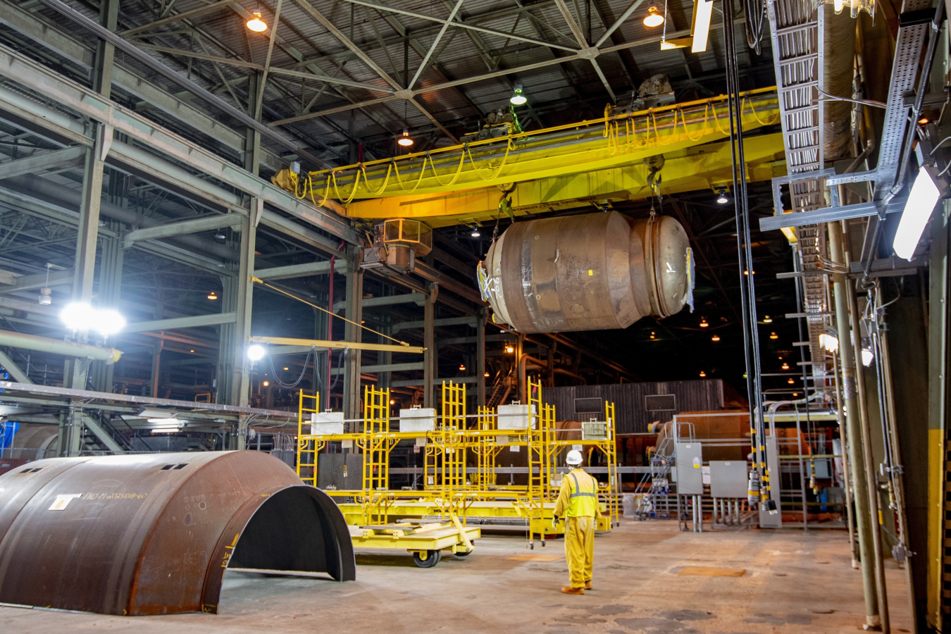 The inside of a large waste facility with lots of pipes and large brown converters. One converter is on the ground and the other is being lifted by a machine 