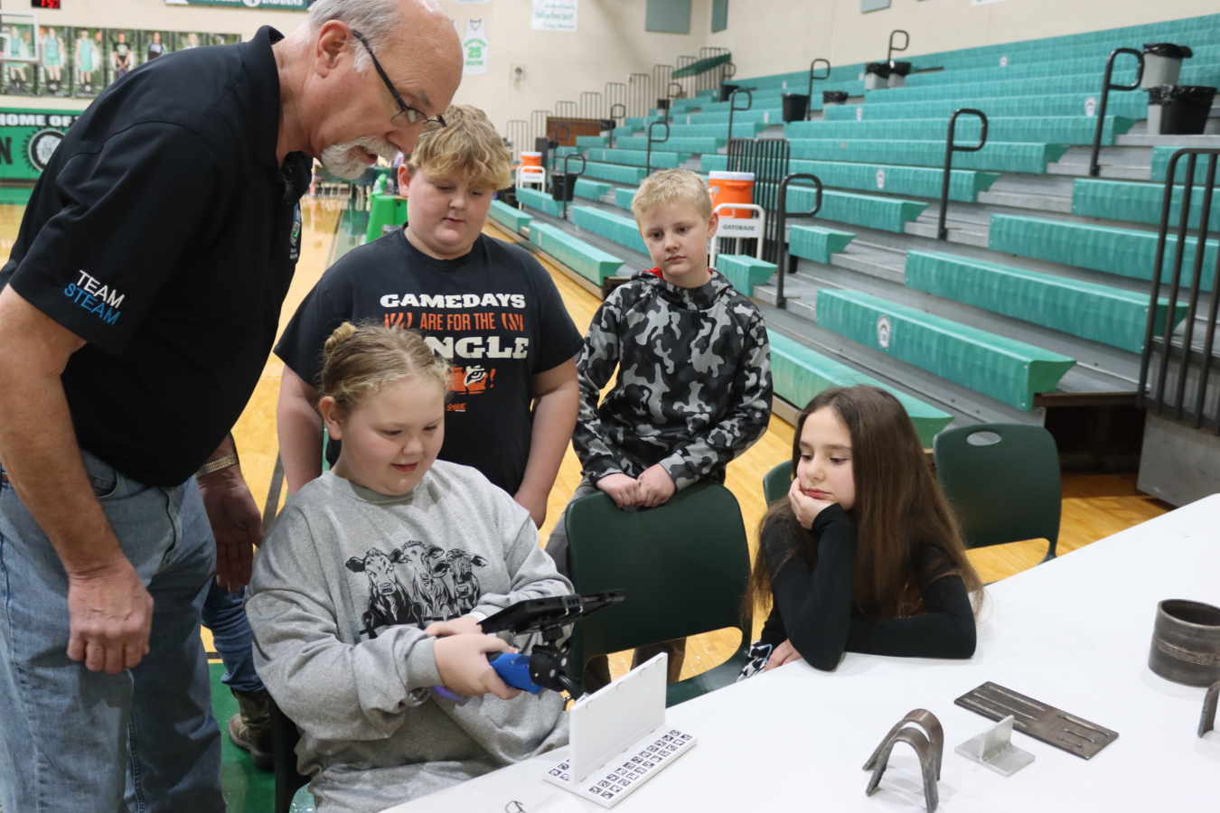 Two elementary school students sit at a table and participate in a virtual welding activity, a man stands behind them helping them out, along with two other students