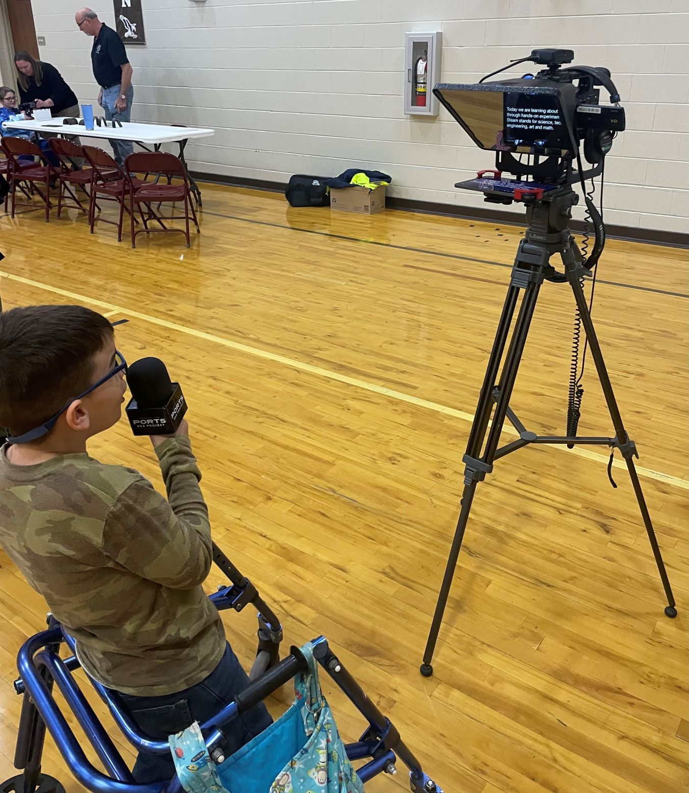 An elementary school student faces a camera and reads off a teleprompter into a microphone