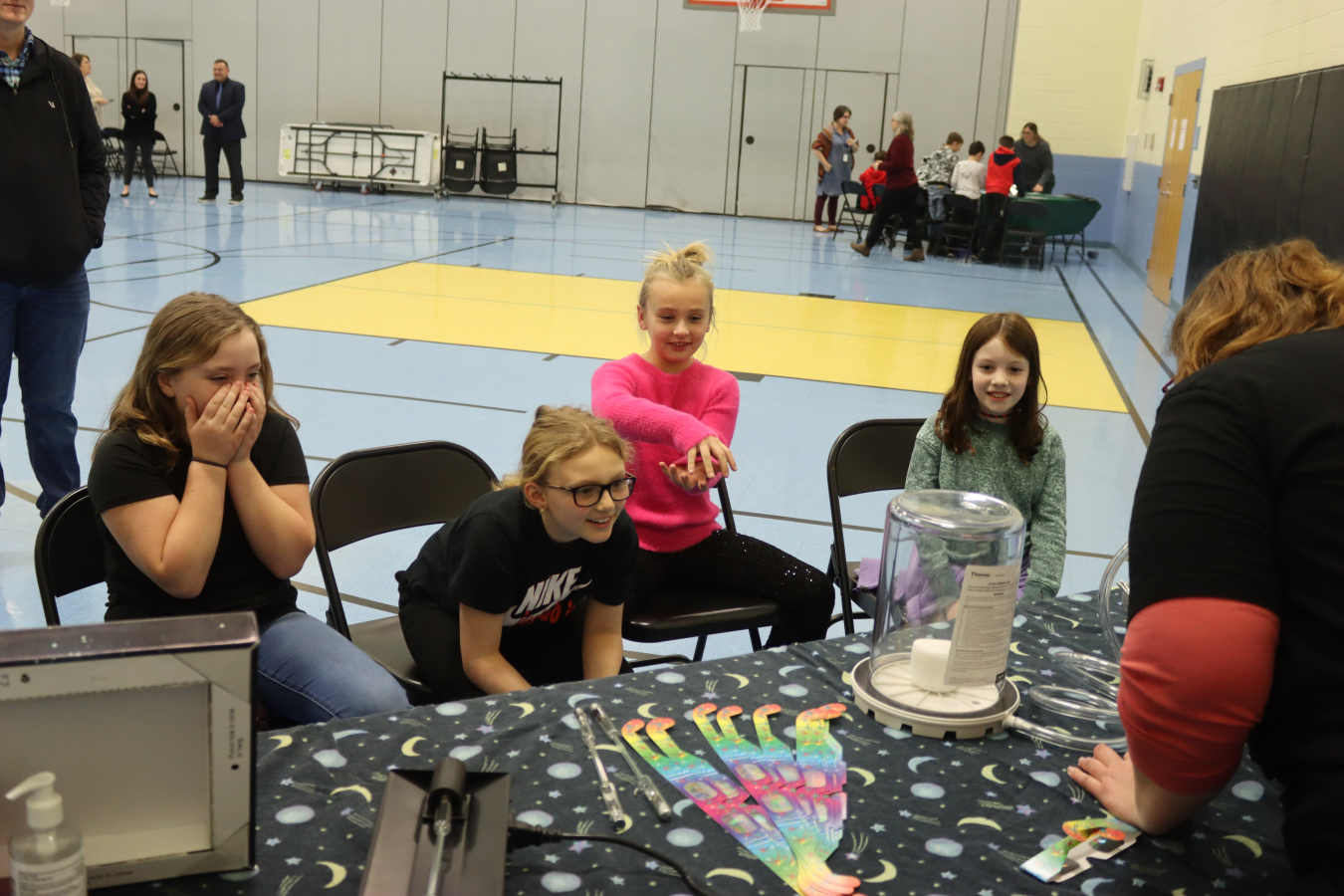 Four elementary school students sit in chairs around a table and watch a science demonstration be performed
