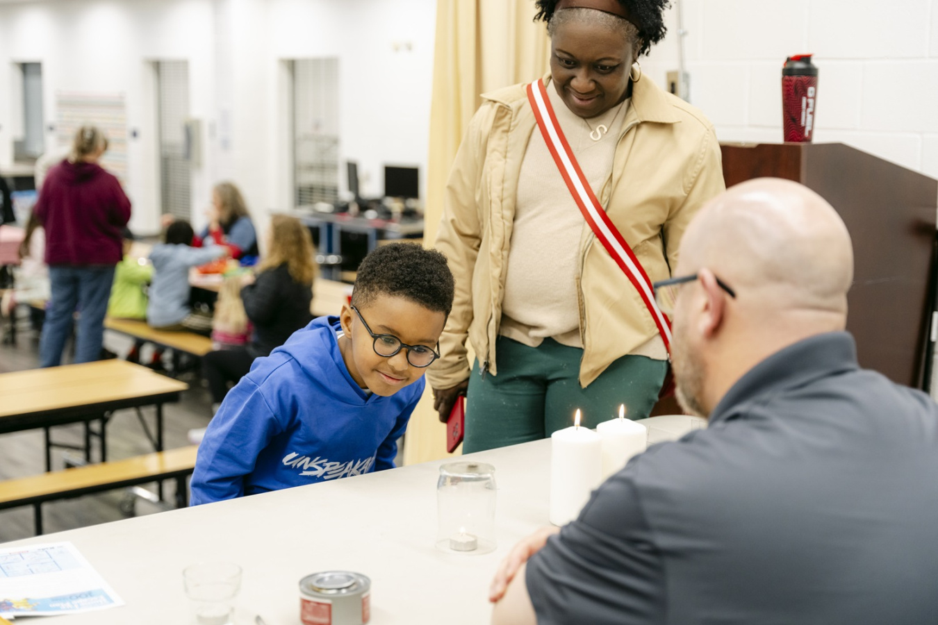 A man with his back turned to the camera sits at a table with a young boy while his mother stands behind him