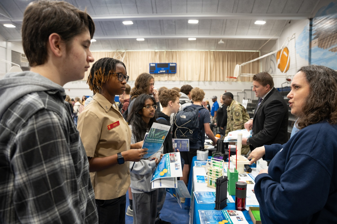 Lab employees stand at a booth at a STEM event and speak to multiple high school students