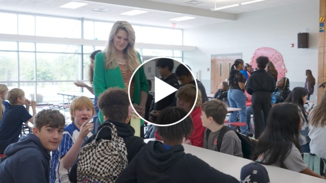 Play button over a still screen of a YouTube video of a woman standing beside a table of middle school students, she talks with them 