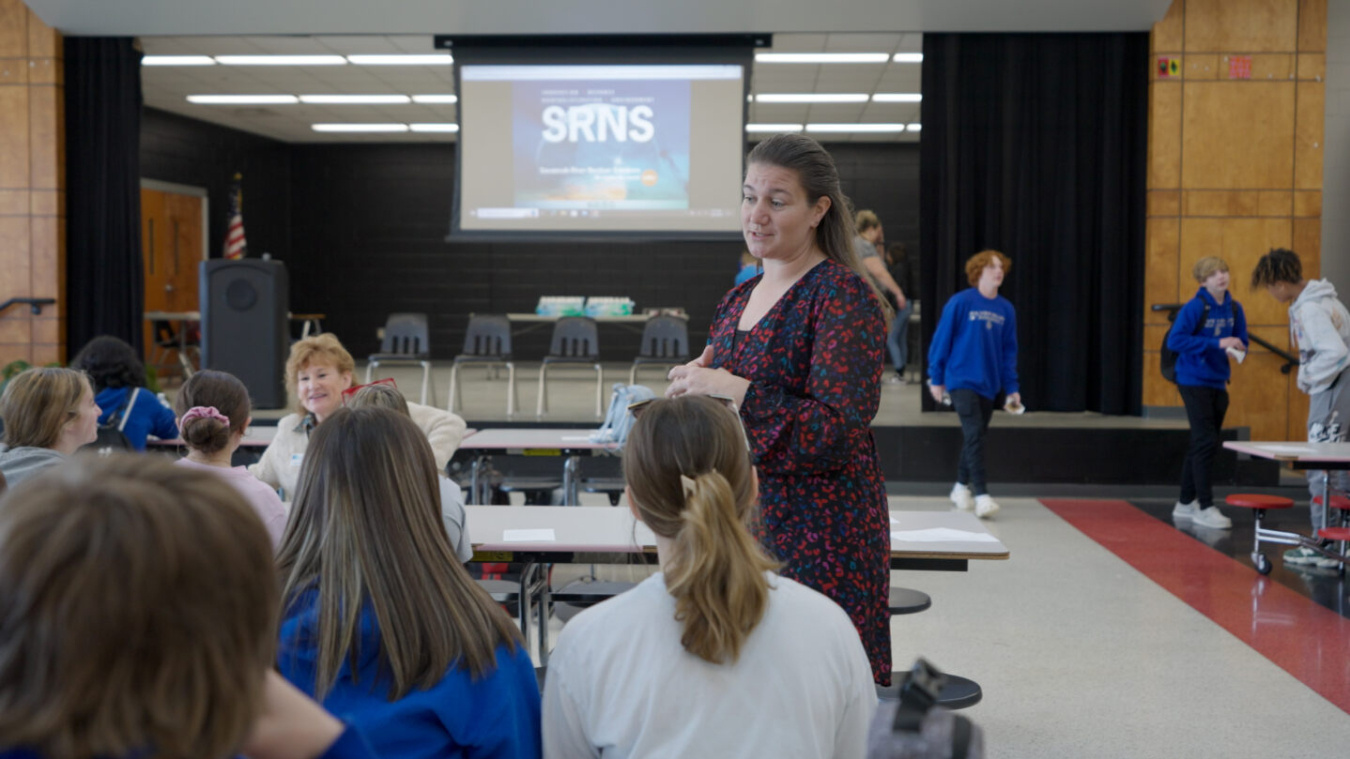 A woman stands and speaks to middle school students sitting at a lunch table