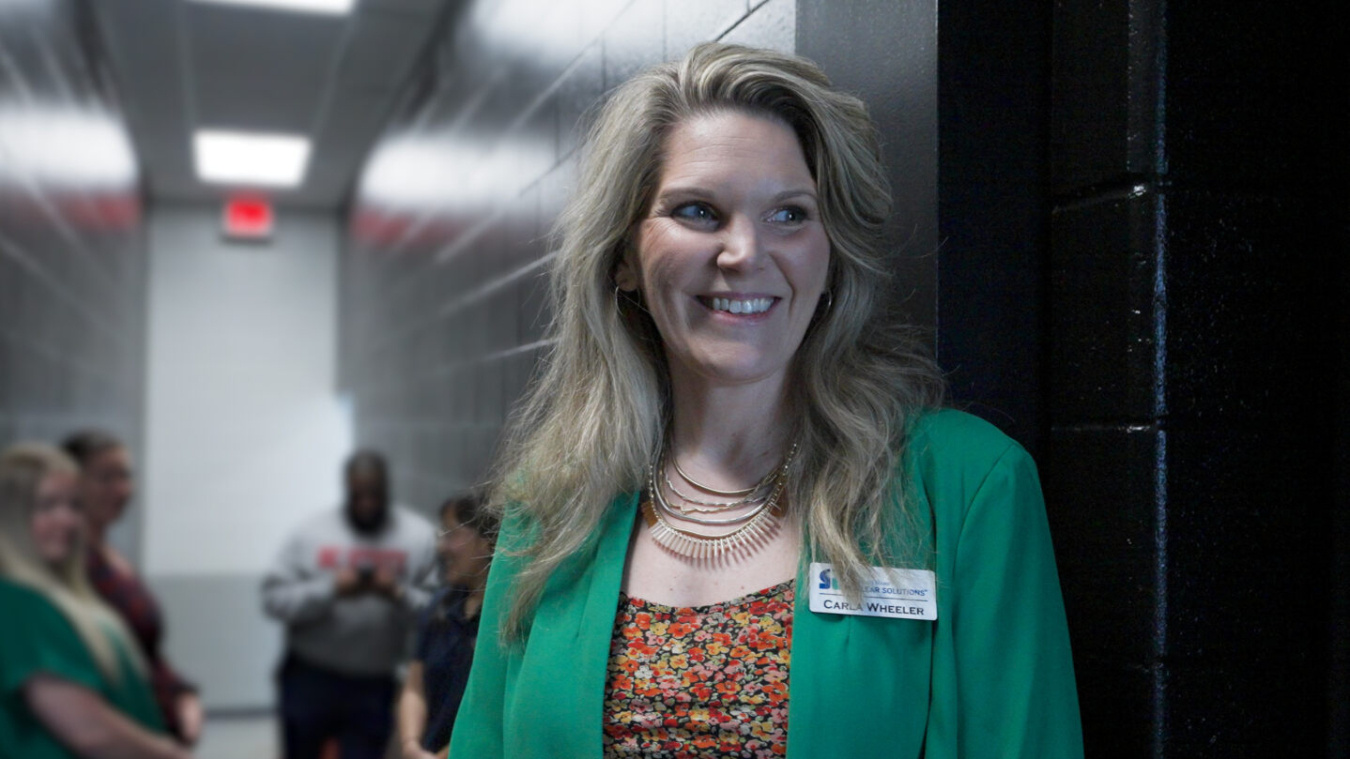 Headshot of a blonde woman with a green jacket and floral top. She stands against a black wall
