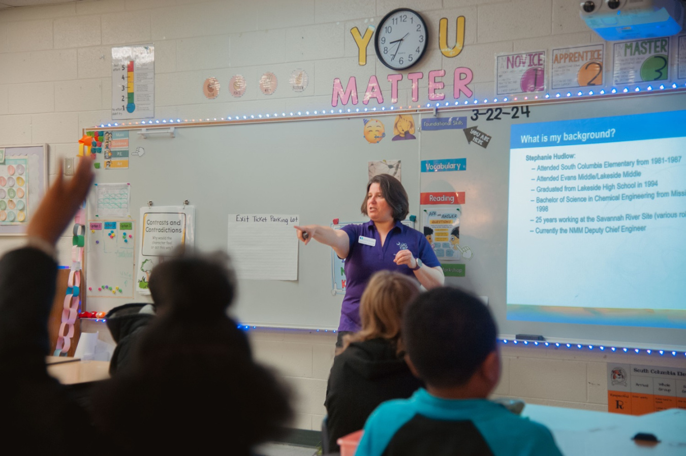 A woman stands at the front of a classroom with a powerpoint presentation beside her, she speaks to a classroom full of students