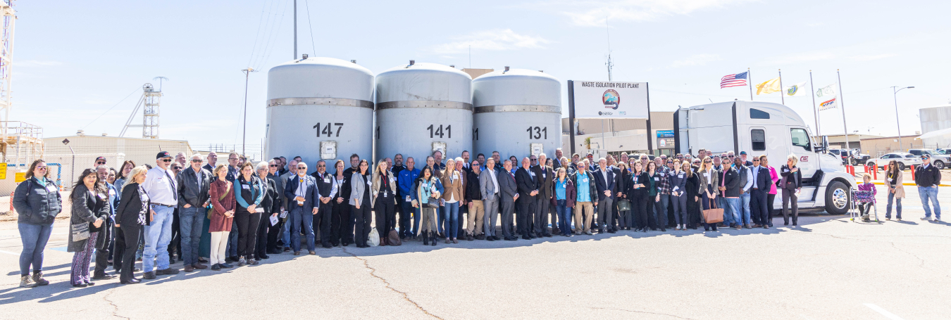 A large group of people pose for a picture in a parking lot standing in front of a truck carrying 3 large, grey canisters 
