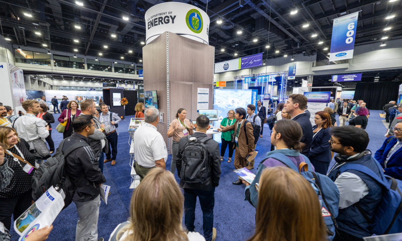 A group of people stands around a speaker at a booth on a convention trade show floor.