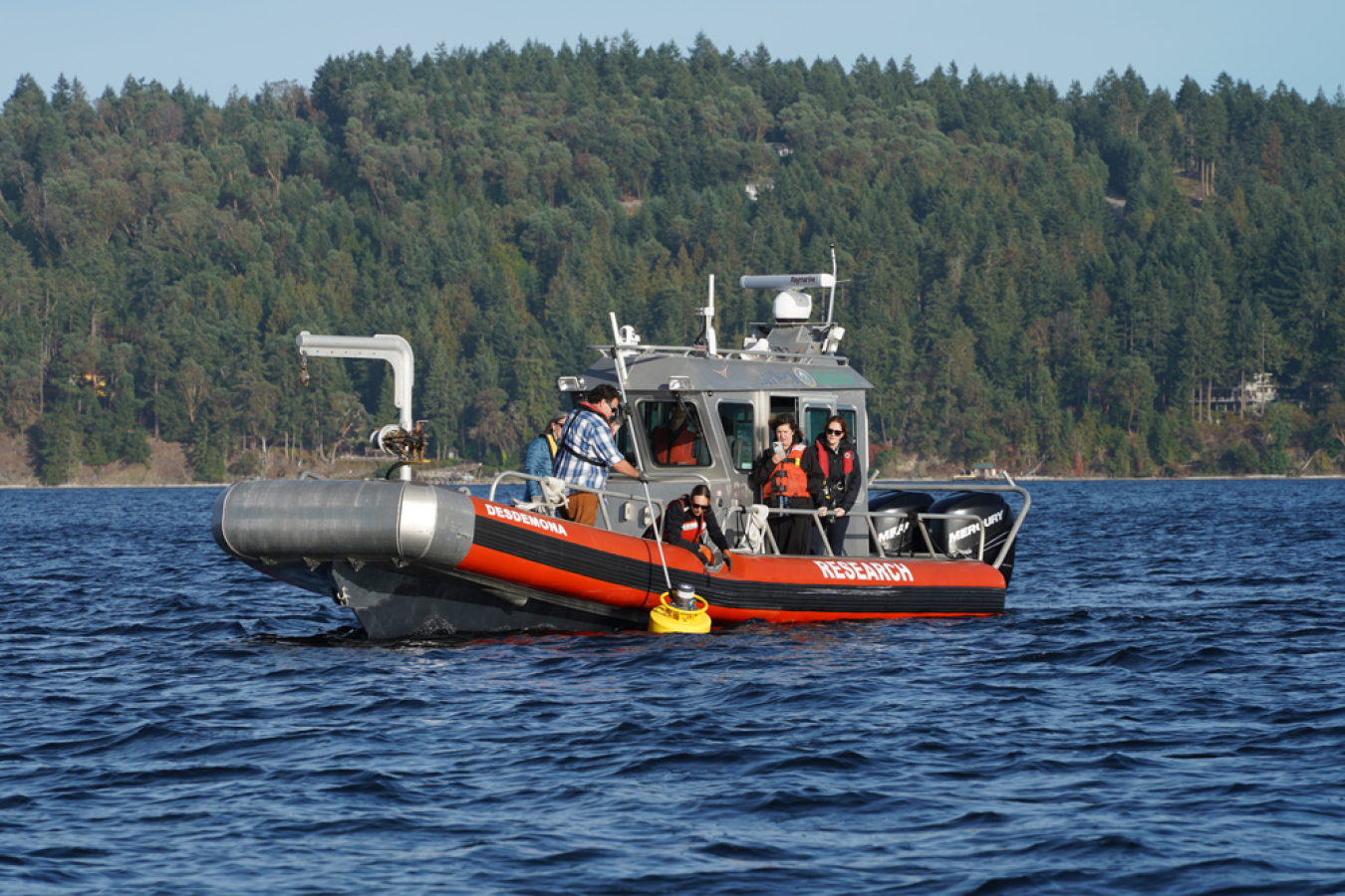 A group of people stand on a boat. One person is using a pole-like device to maneuver a yellow device in the water.
