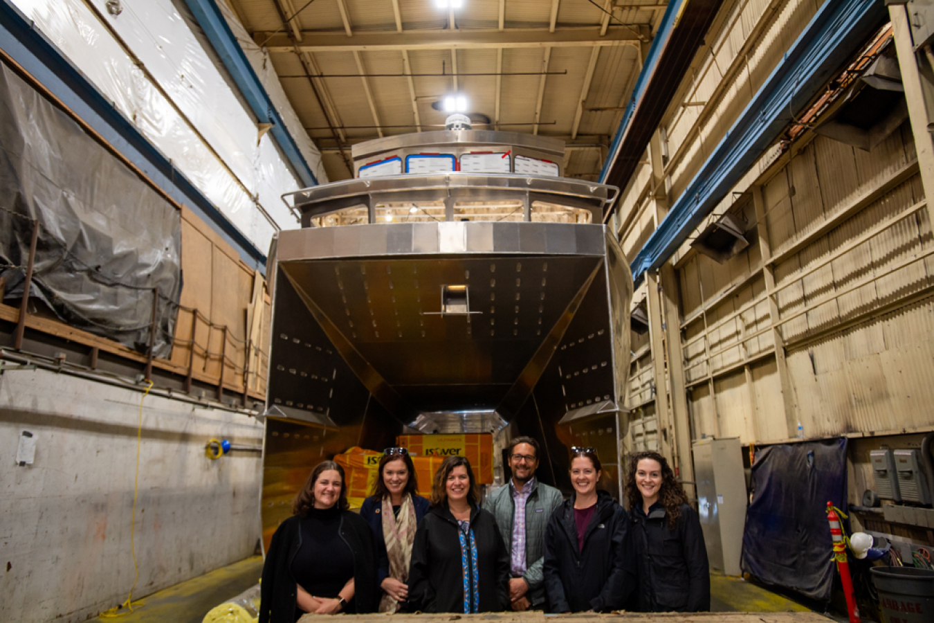 Six people stand in front of a partially constructed boat.
