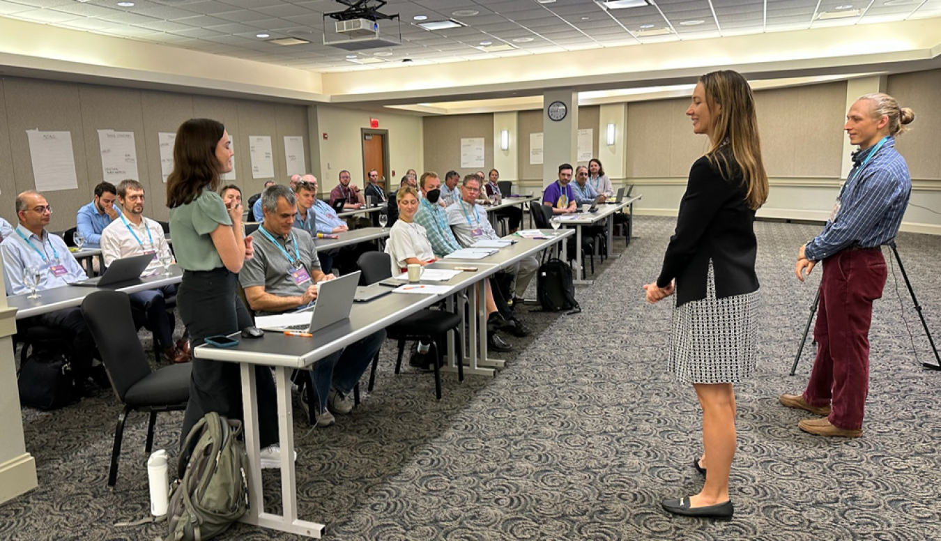 Two people are standing in front of a conference room facing people sitting at several rows of tables. Another person standing at one of the tables is speaking to the group.