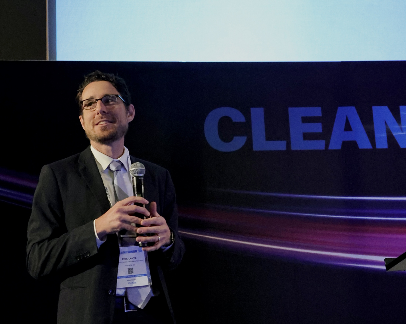 Man with curly hair and glasses in a black suit stands holding a mic in front of a backdrop that says "CLEAN".
