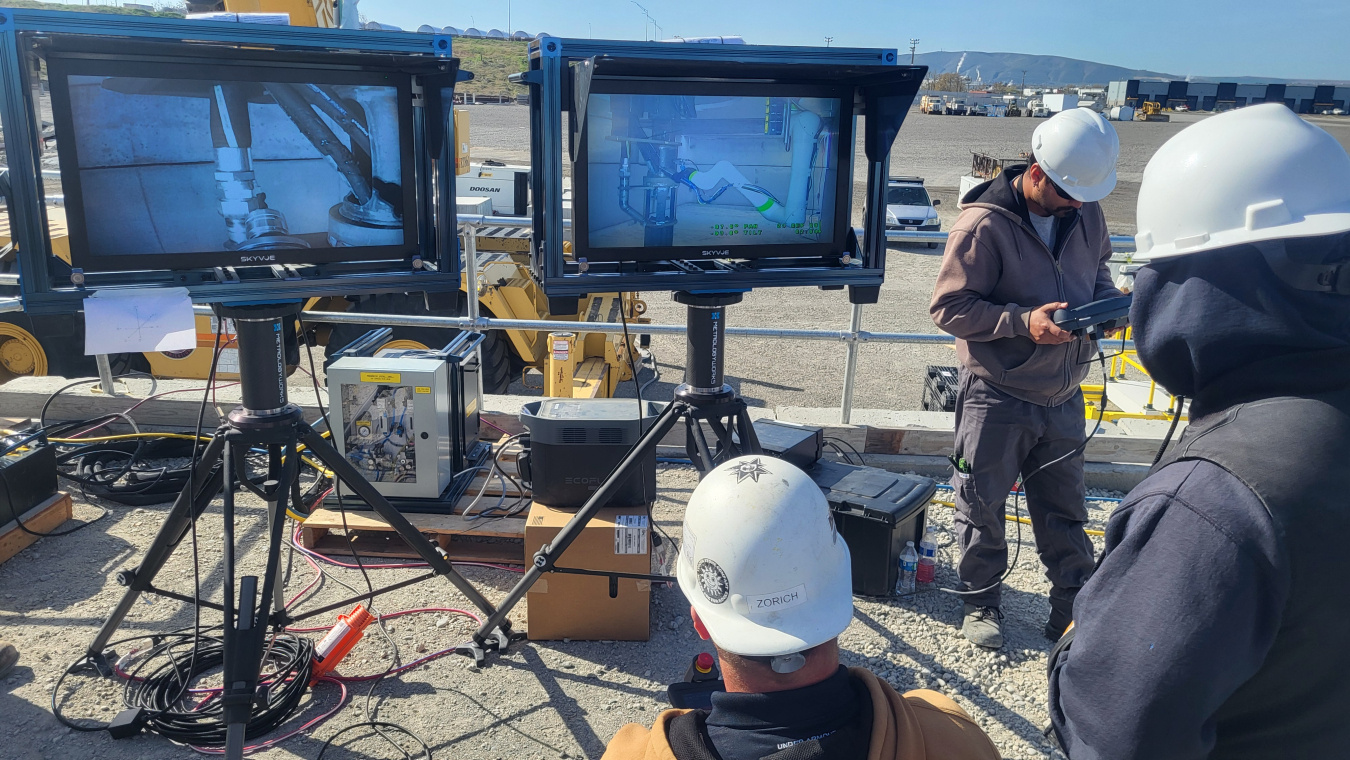 Two large screens displaying a machine. 3 workers with white construction hats stand around the screens