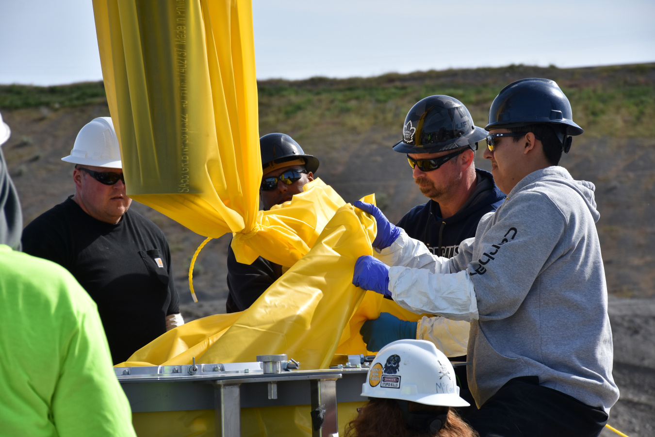 Men in construction hats stand around a tall yellow cylinder, using it to demonstrate a process