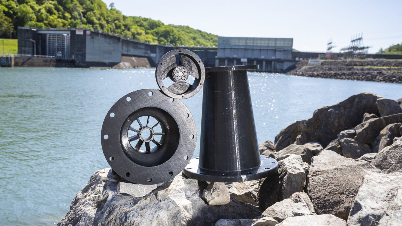 Three hydropower components sitting on rocks along a waterway with a hydropower facility in the background.