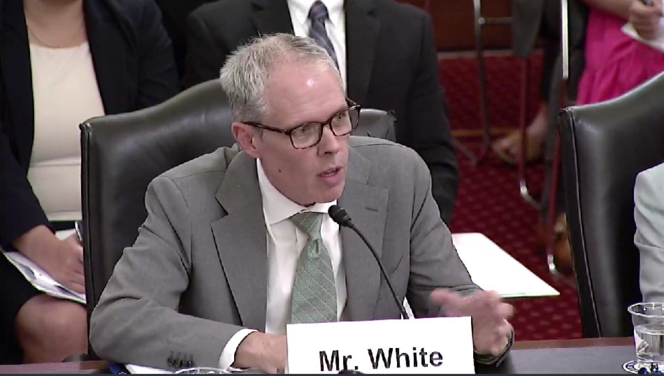 A man in a grey suit and glasses sits at a table with a name tag in front of him that reads "Mr. White"