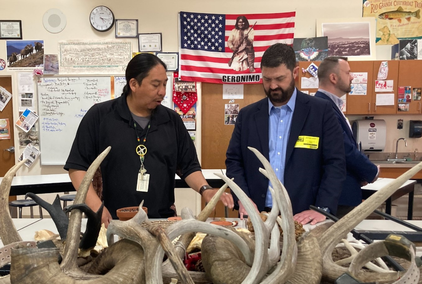 Two men stand in front of a table with antlers on it while speaking to each other