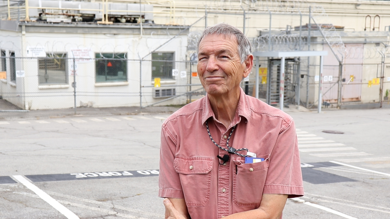 A man in a light pink shirt in the middle of the frame with a factory building background behind him