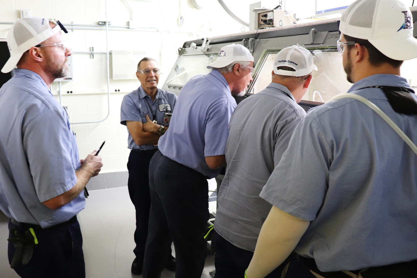 A group of employees working with a glovebox wearing white hats and blue shirts