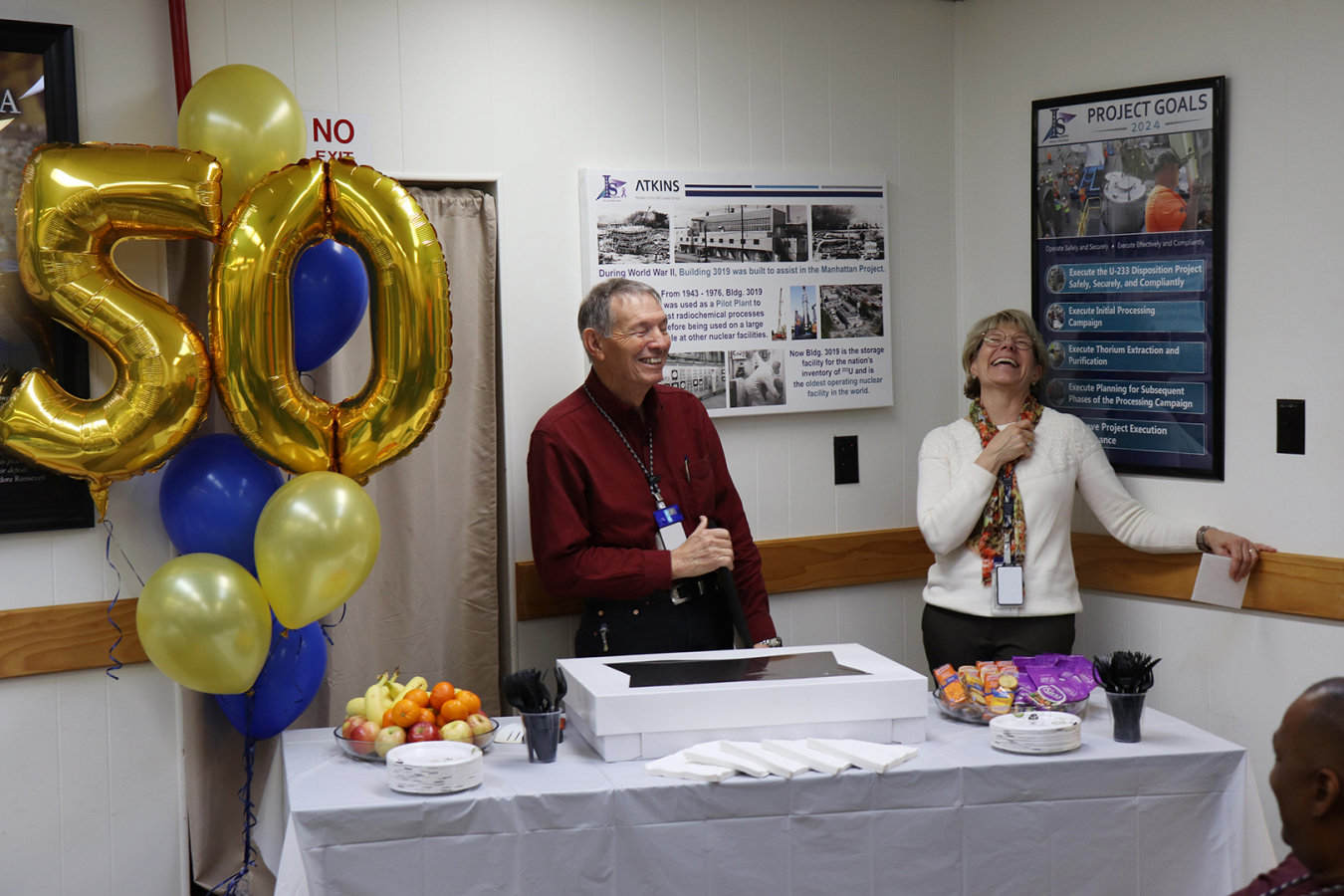 A man and a woman stand behind a table with a cake on it beside gold balloons of the number 50