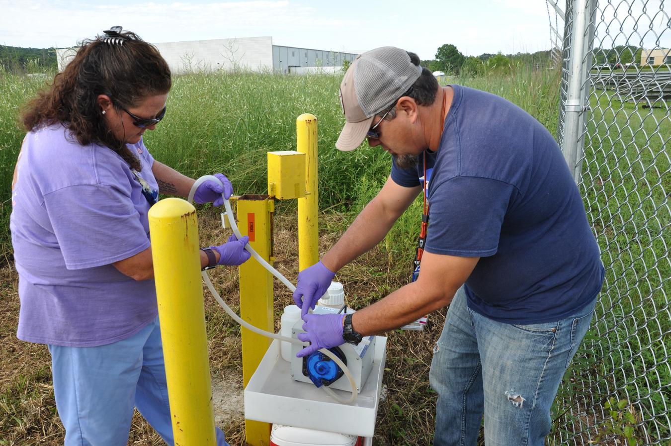 Two employees work on a sampling system outside between yellow poles