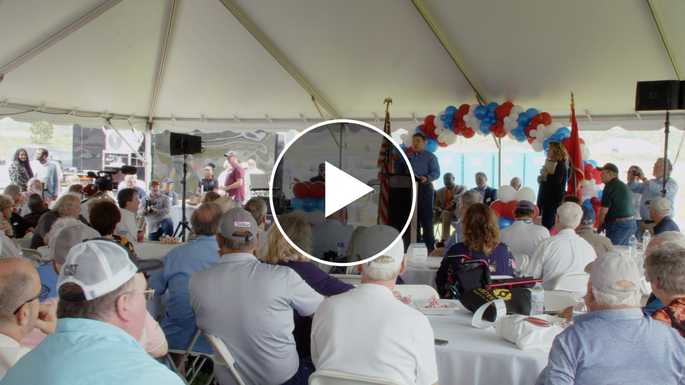 Still from a video of a large group of people in a white, outdoor tent. There is a play button in the middle of the picture