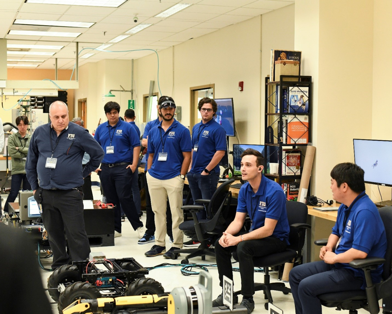 A group of college students in blue shirts sit and stand around a room