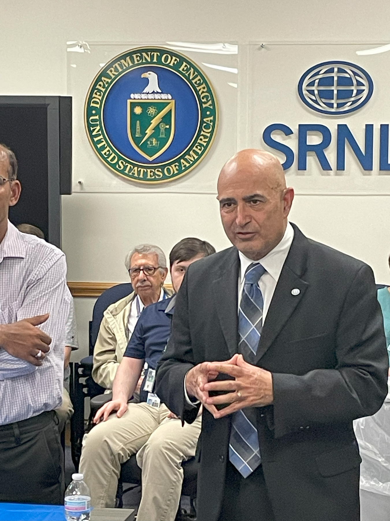 A man speaking in the front of a room with other participants and the DOE seal behind him
