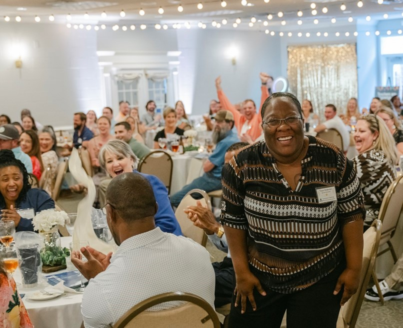 A smiling woman stands up from a table while the room of people applaud her