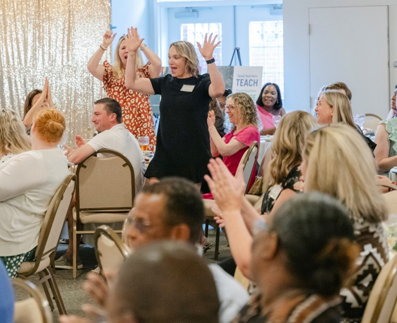 A woman with both her hands up stands up from a table while the room of people applaud her