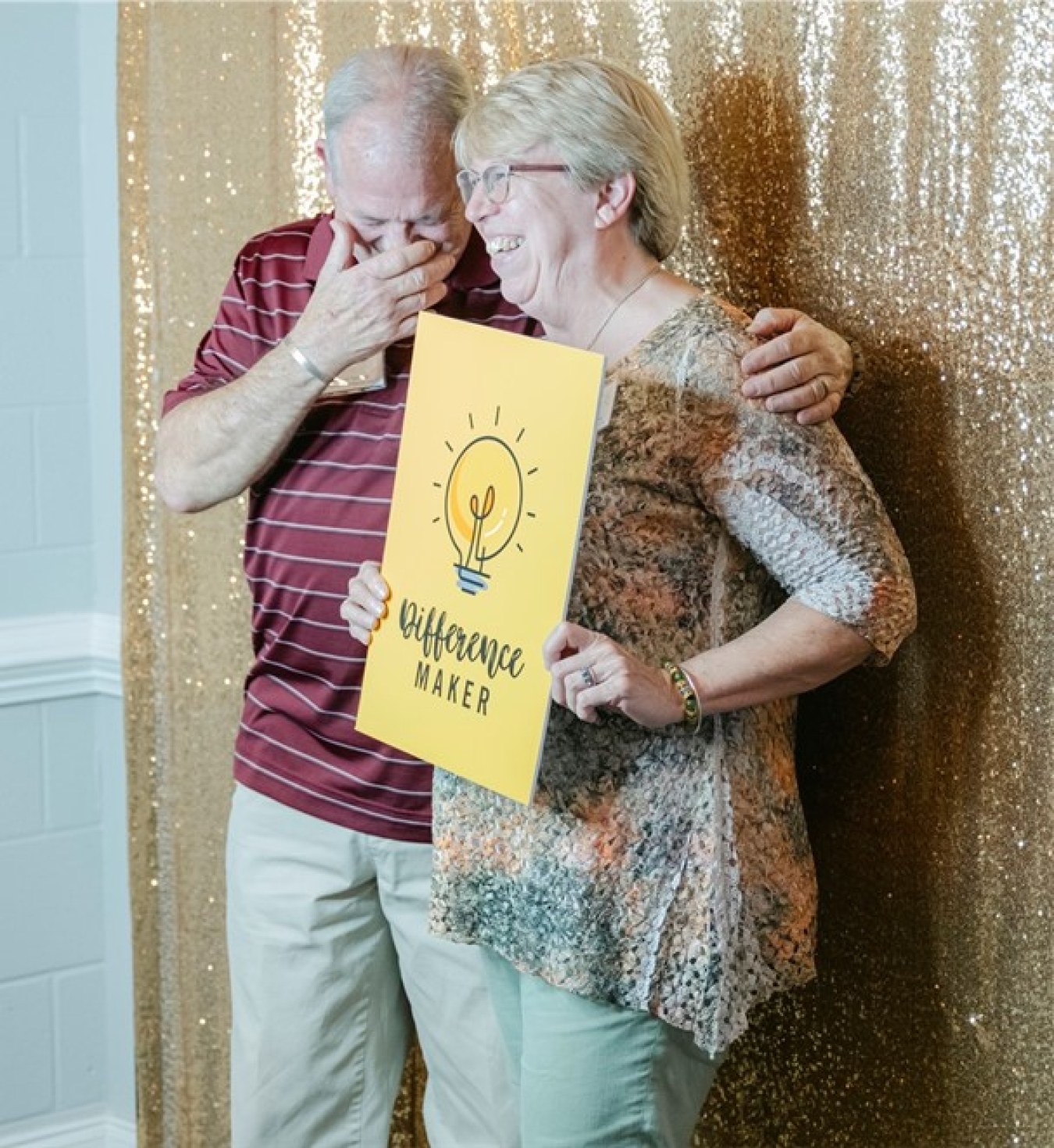 A smiling man and woman pose for a picture while holding a yellow sign that reads "Difference Makers"