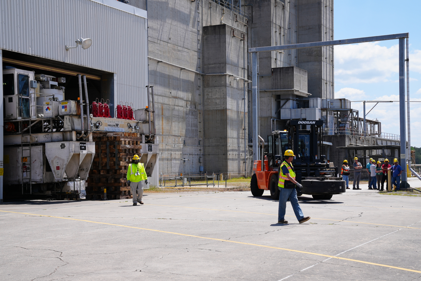 Employees in yellow hard hats and vests work beside a building with large construction vehicles 