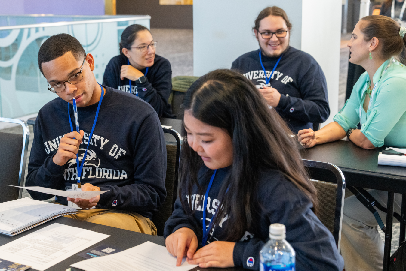 Two students sitting at a table in the foreground. Three people are talking at another table behind them.