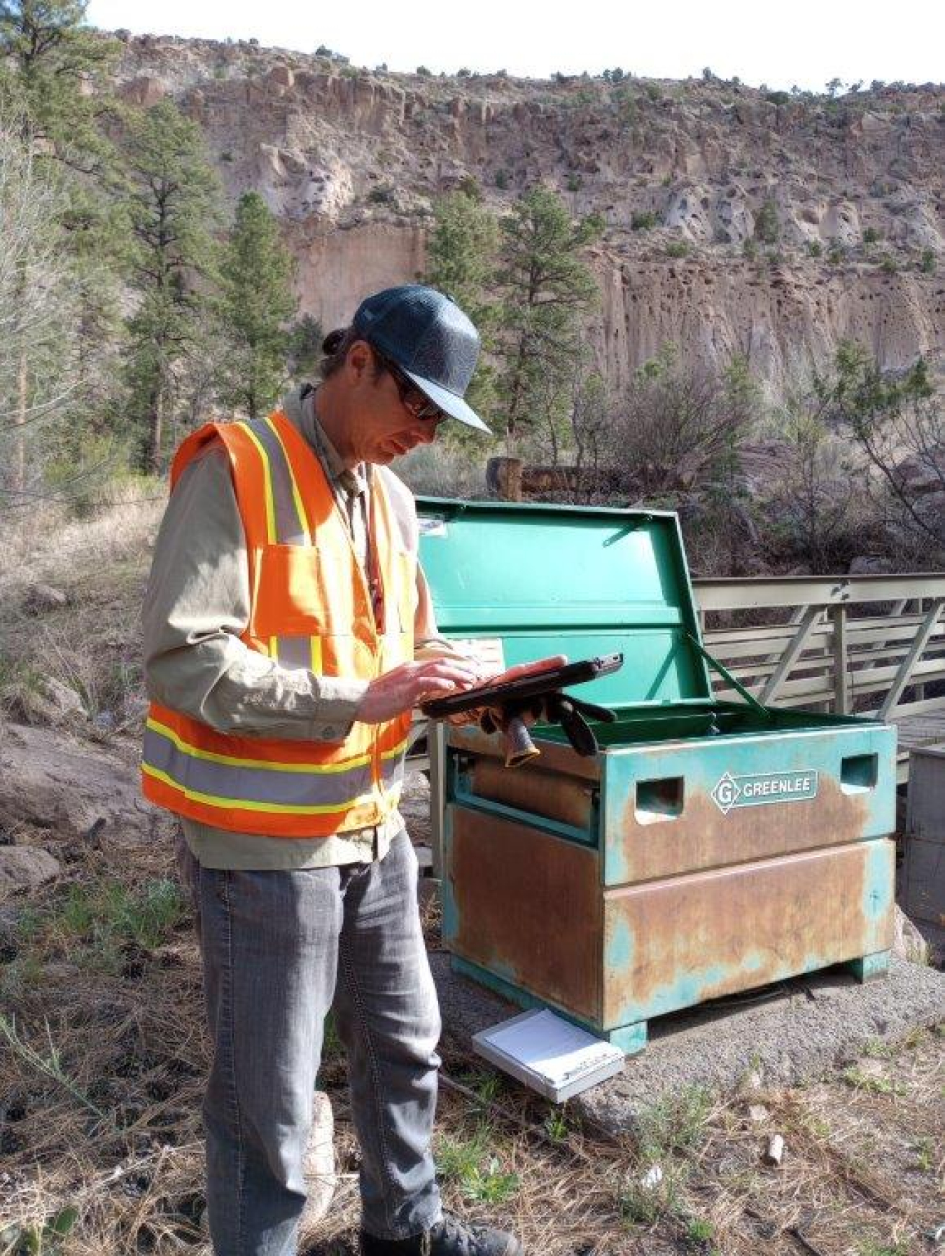 A man in an orange safety vest and hard hat stands and uses a device in a forest area