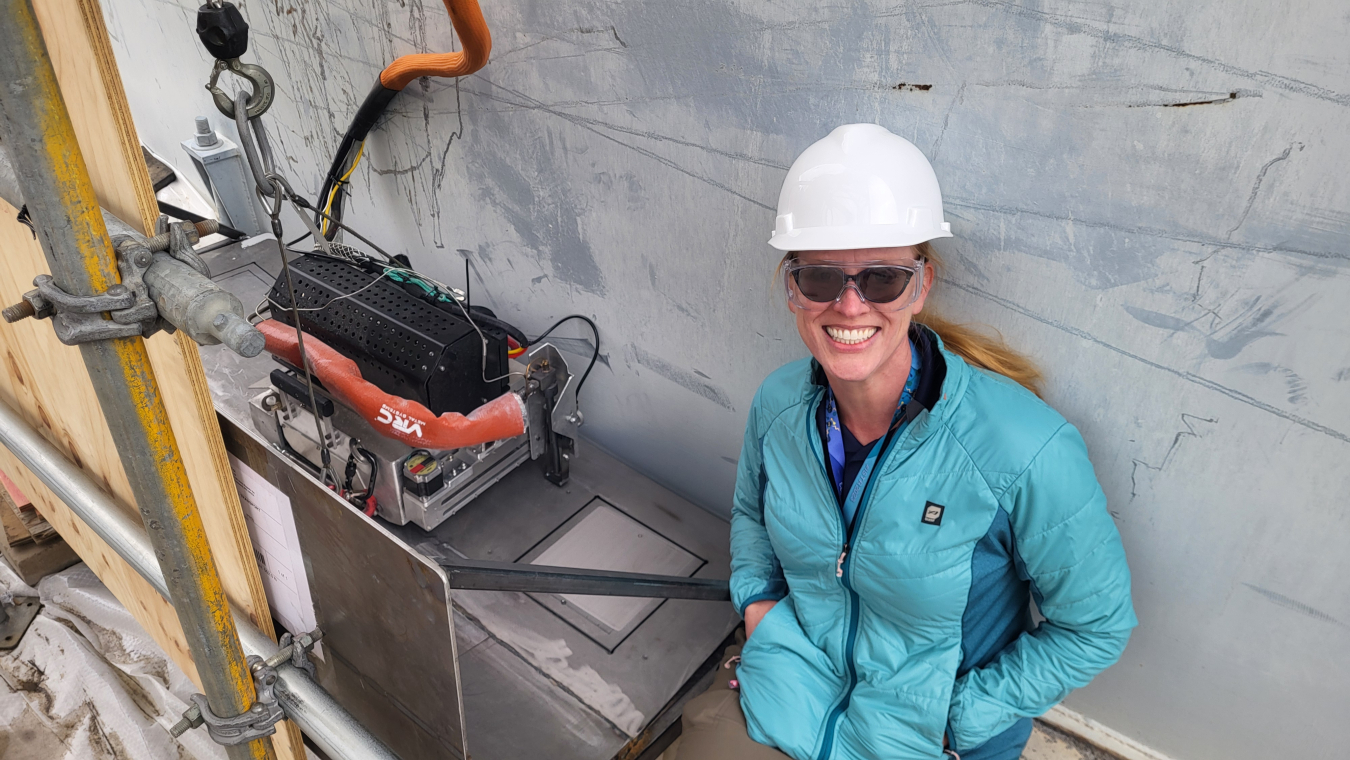 A woman in a white hard hat and a blue jacket sits against a wall and poses next to a metal patch being applied to the wall beside her