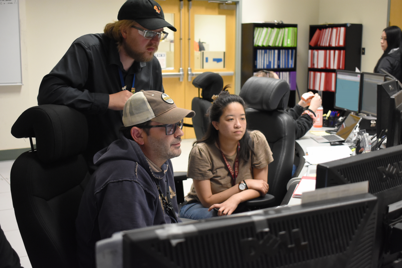 A group of people sit and stand around a computer and look at the computer screen