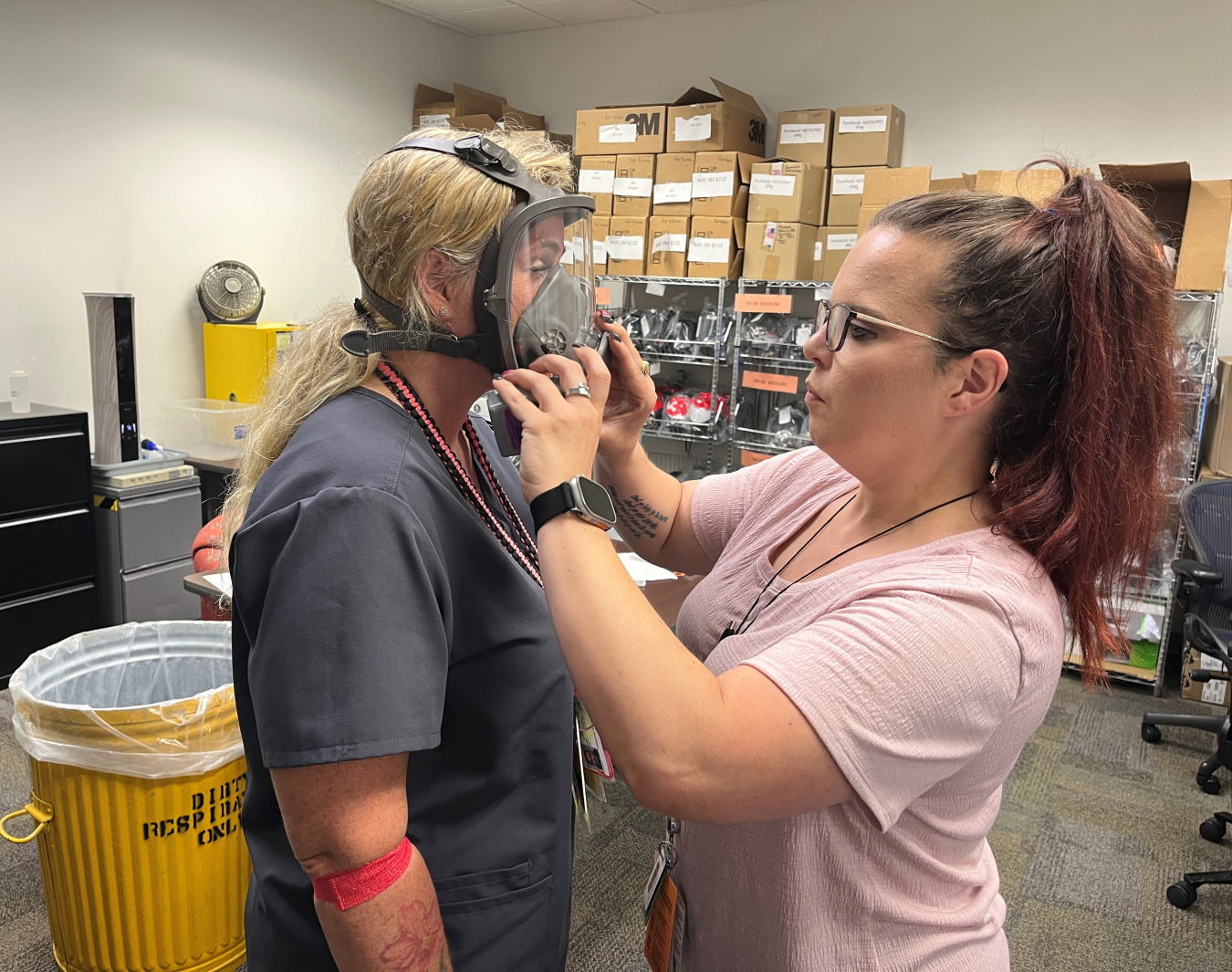 A woman puts a respirator onto another woman and tests to see if it fits
