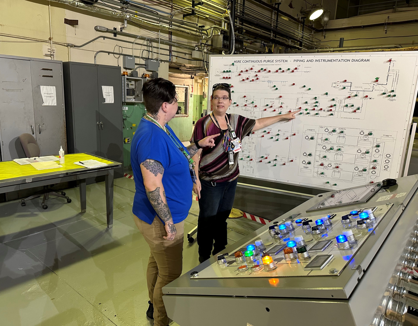 Two women stand in front of a large control board and a whiteboard on wheels, one woman points to the whiteboard while the other looks at her