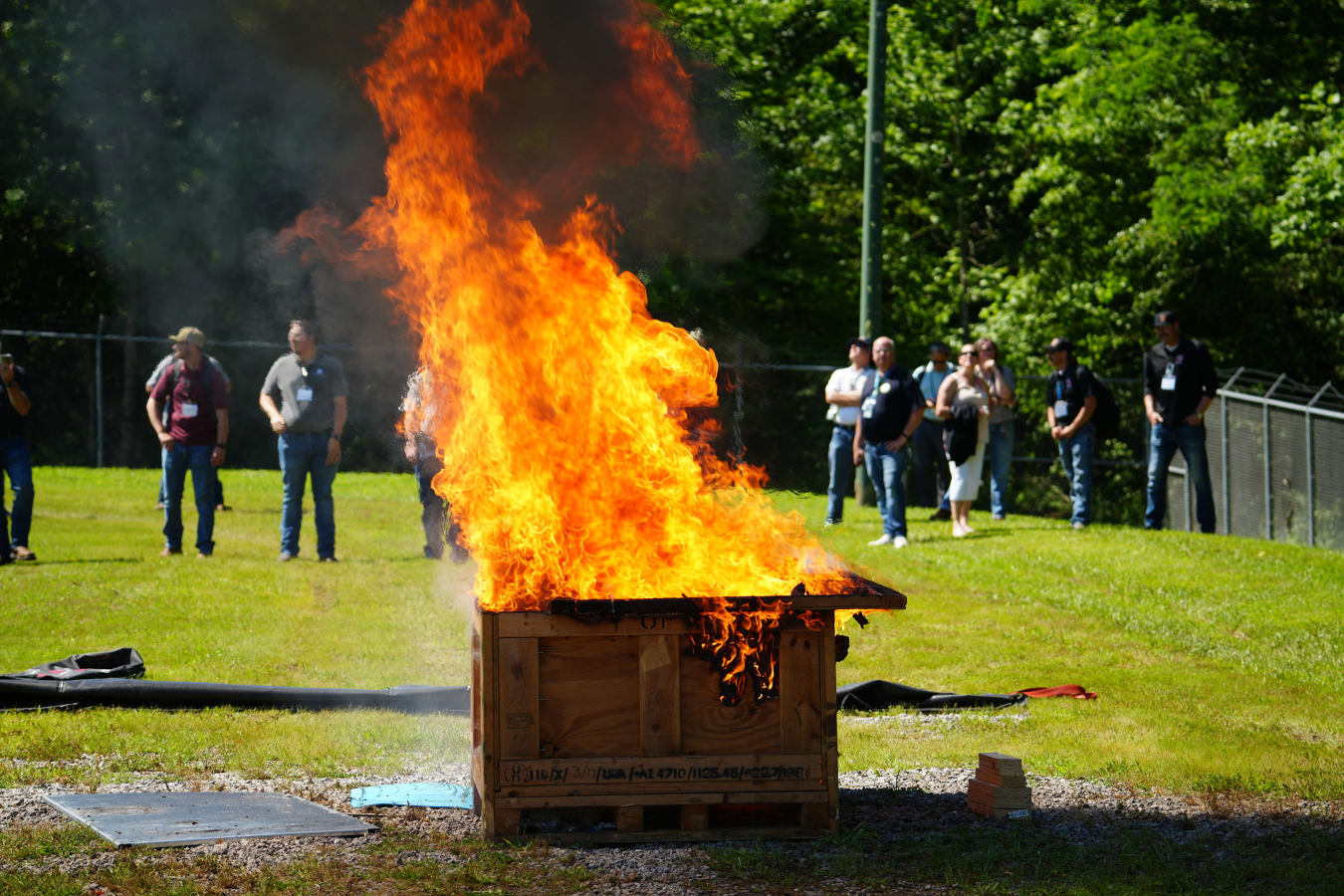A fire burns inside and on top of a large wooden box, workers stand around the box and observe