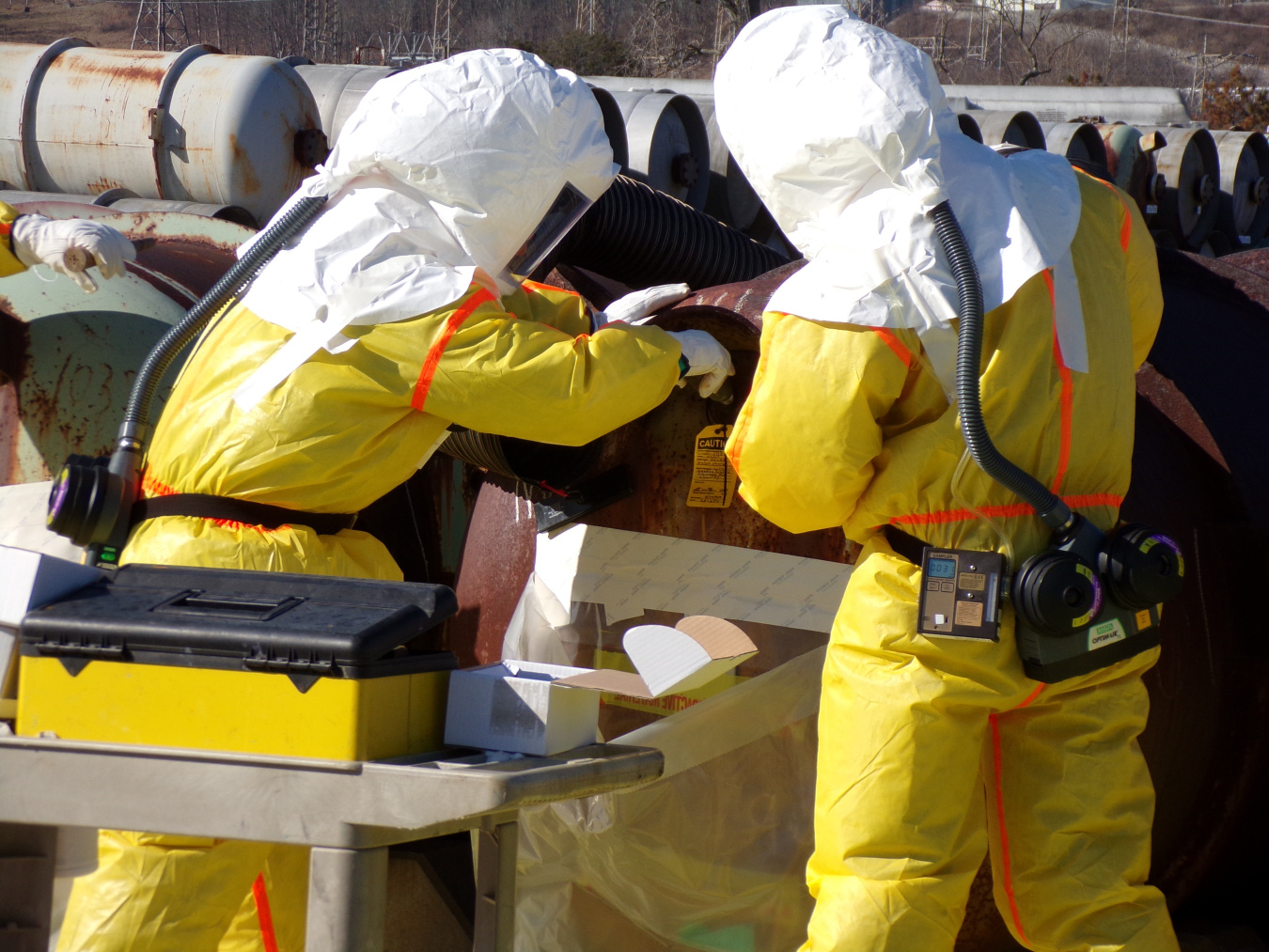 Two employees in yellow and white hazmat suits work on pipes with their backs turned towards the camera