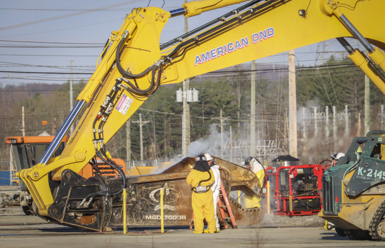 A large yellow construction crane vehicle works with employees standing around it in yellow suits