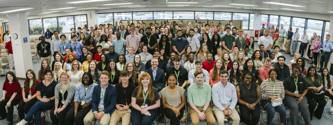A large group of young interns sit and stand in a room and pose for a group photo