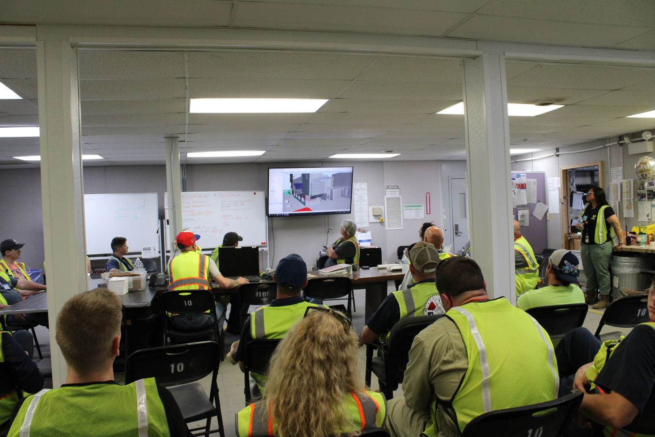 A group of adults sit in a classroom and watch a model of a building being displayed on a TV in the front of the room