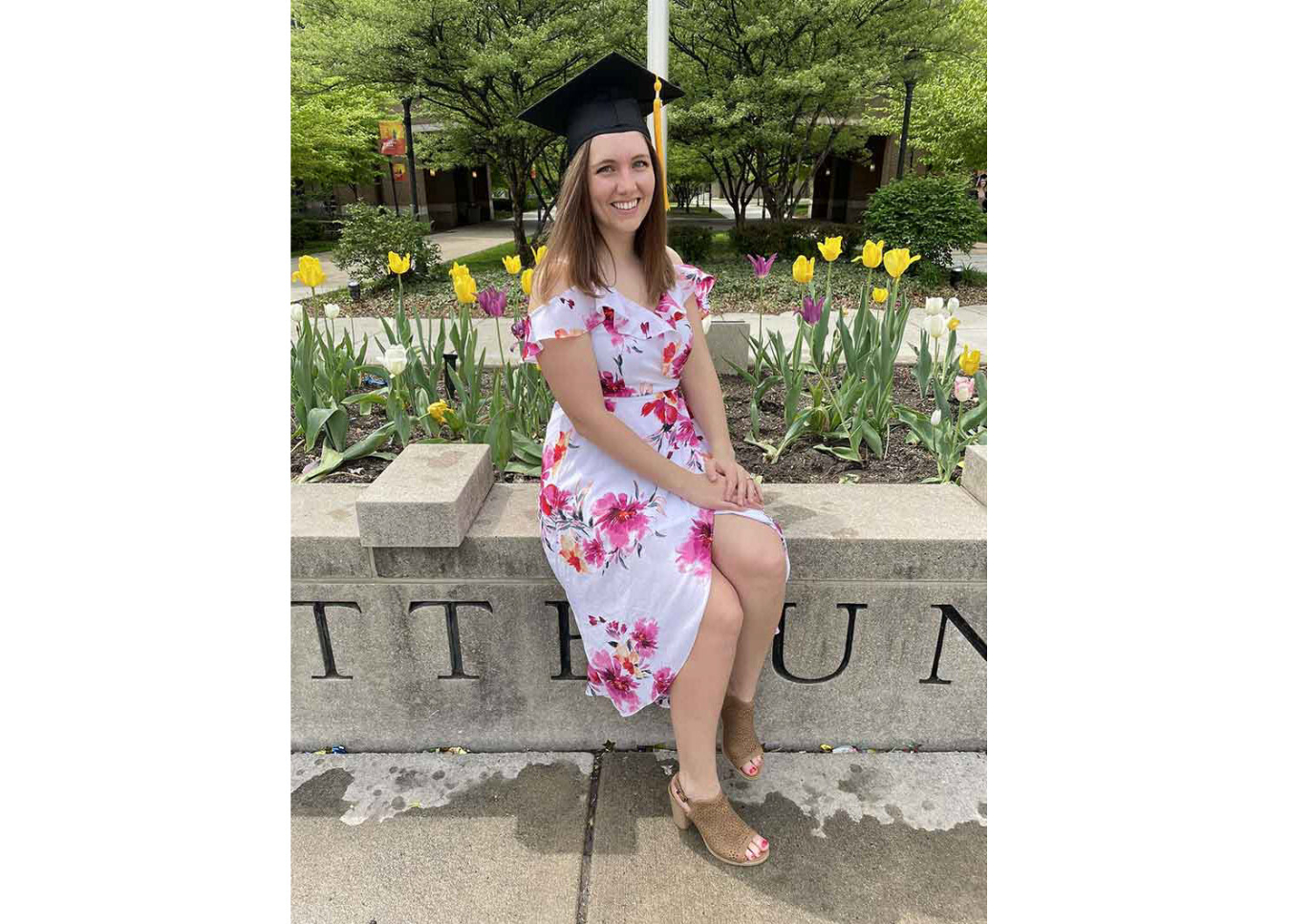 Emily Corcoran sitting in front of a flower bed filled with tulips wearing a floral dress and graduation cap.